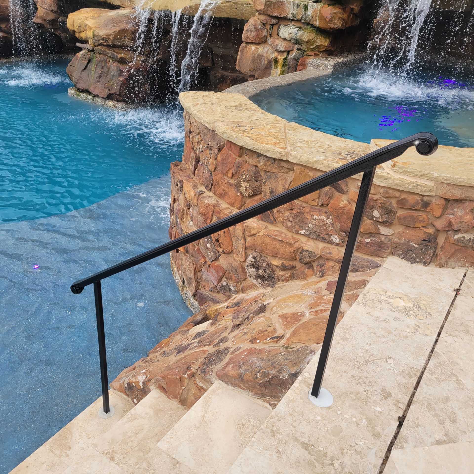 Black ornamental metal handrail beside a backyard pool and stone waterfall feature